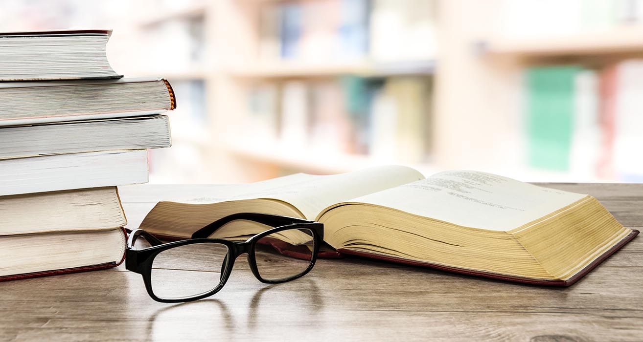 A pair of glasses rest in front of a stack of books on a desk.