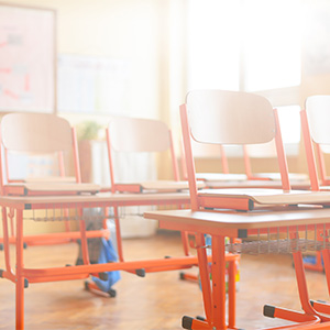 Two rows of student desks and chairs sit empty in a sunny classroom.