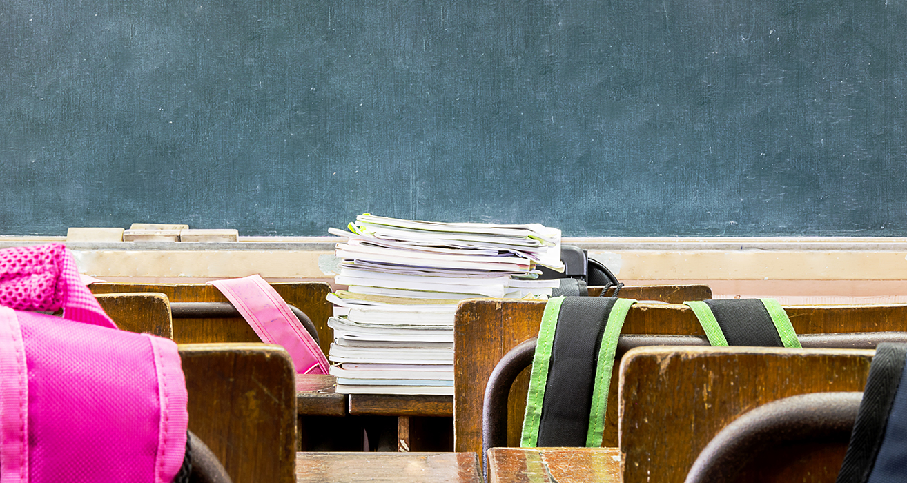 Chairs with backpacks on them face a teacher’s desk piled with notebooks.